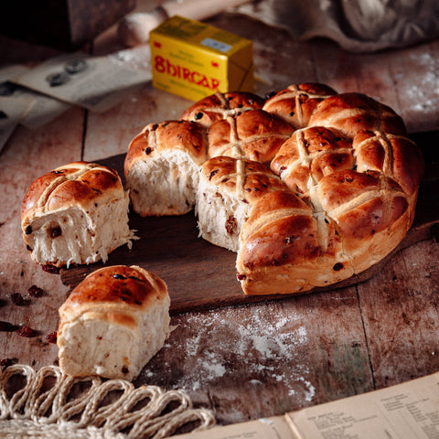 A freshly baked loaf of hot cross buns with one piece torn away, revealing the studded dried fruits and cross design. Set on a rustic wooden table with a newspaper and a box of tea in the background.